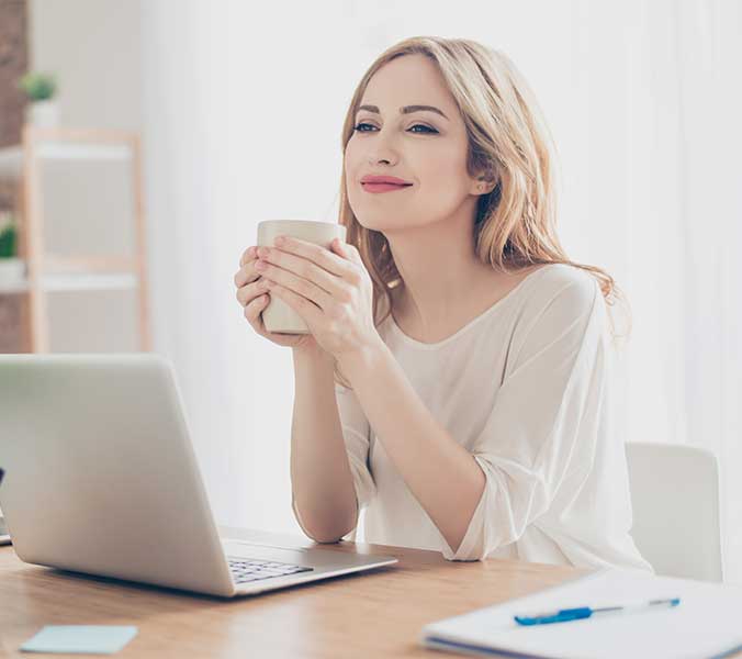 Daydreaming woman sitting at a desk with a laptop while holding a cup.